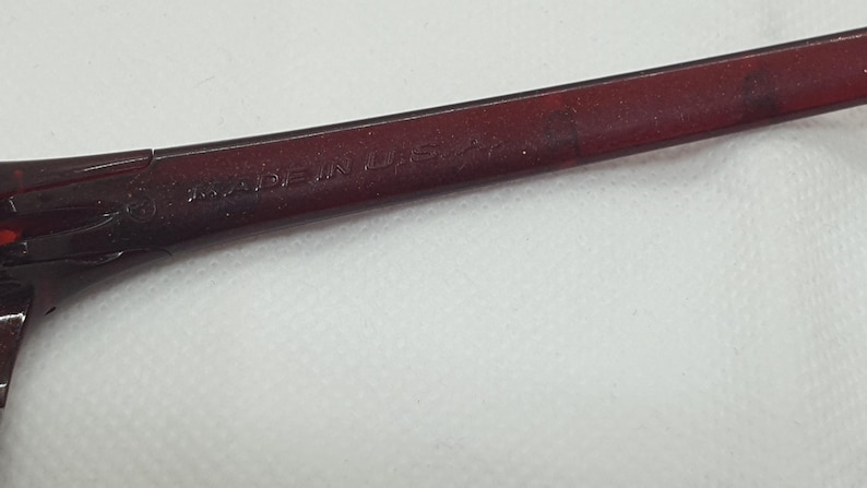 May include: Close-up of a section of eyeglasses. The frame is a dark burgundy colour. The text "MADE IN USA" is visible on the frame.