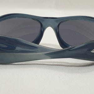 May include: Dark blue sunglasses with gray lenses. The sunglasses have a curved frame and arms, with a silver logo on the side. The lenses are a dark gray color, and the frame is a translucent blue.