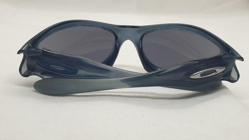 May include: Dark blue sunglasses with gray lenses. The sunglasses have a curved frame and arms, with a silver logo on the side. The lenses are a dark gray color, and the frame is a translucent blue.