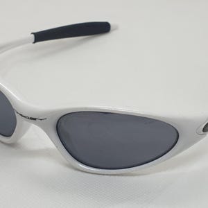 Puede incluir: Gafas de sol blancas con lentes grises y puntas de patillas negras. Las gafas tienen una montura curvada y un dise&ntilde;o deportivo. El logotipo de Oakley es visible en la patilla. Adecuadas para actividades al aire libre.
