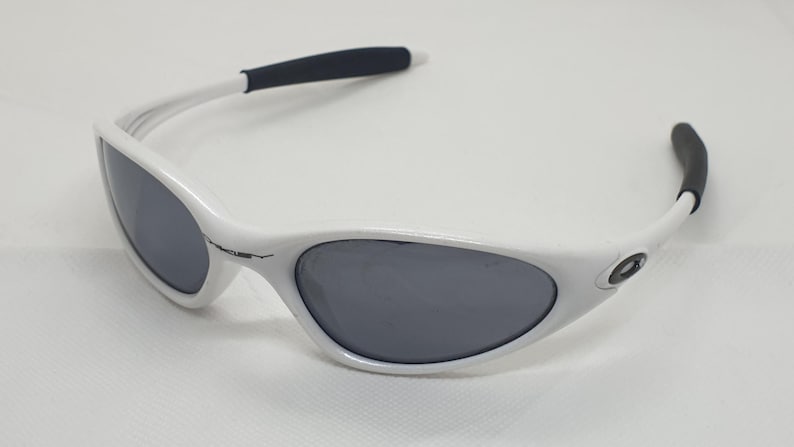 Puede incluir: Gafas de sol blancas con lentes grises y puntas de patillas negras. Las gafas tienen una montura curvada y un dise&ntilde;o deportivo. El logotipo de Oakley es visible en la patilla. Adecuadas para actividades al aire libre.