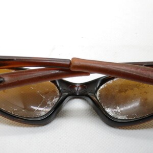 Puede incluir: Gafas de sol marrones y negras con un dise&ntilde;o de montura &uacute;nico. Las lentes son de color marr&oacute;n claro. Las patillas de las gafas de sol est&aacute;n plegadas hacia adentro. Las gafas de sol est&aacute;n sobre un fondo blanco.
