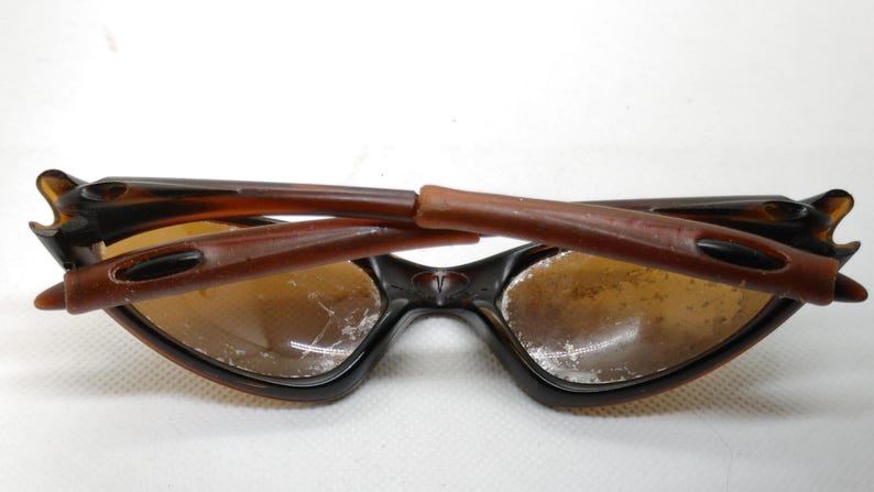 Puede incluir: Gafas de sol marrones y negras con un dise&ntilde;o de montura &uacute;nico. Las lentes son de color marr&oacute;n claro. Las patillas de las gafas de sol est&aacute;n plegadas hacia adentro. Las gafas de sol est&aacute;n sobre un fondo blanco.