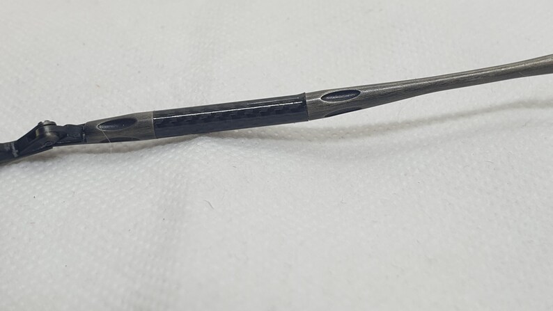 May include: Close-up of a dark gray eyeglass arm with a carbon fiber section. The arm has a sleek, modern design with oval cutouts and a metallic finish. The hinge is visible on the left side.