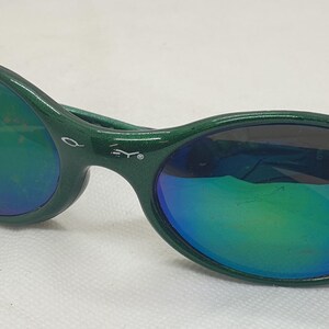 May include: Green sunglasses with iridescent lenses. The sunglasses have a dark green frame and a curved design. The lenses reflect a mix of green, blue, and purple hues. The sunglasses are set against a white background.