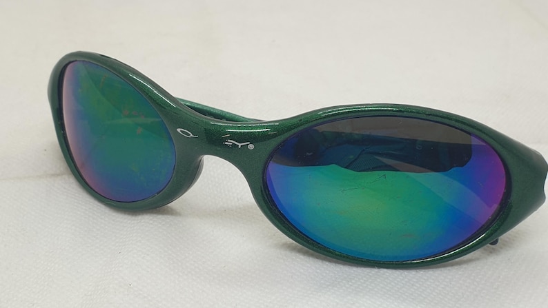 May include: Green sunglasses with iridescent lenses. The sunglasses have a dark green frame and a curved design. The lenses reflect a mix of green, blue, and purple hues. The sunglasses are set against a white background.