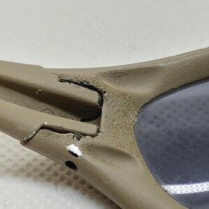Puede incluir: Primer plano de unas gafas de sol. La montura es de color beige con desgaste visible. La lente es de color gris oscuro. El &aacute;rea de la bisagra muestra da&ntilde;os y desconchones de pintura.