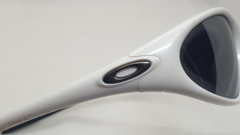 Puede incluir: Gafas de sol blancas con lentes oscuras. La montura presenta un acabado texturizado y un logotipo met&aacute;lico en la patilla. Las gafas de sol tienen un dise&ntilde;o deportivo y moderno.