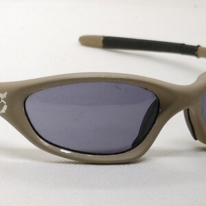 Puede incluir: Gafas de sol color beige con lentes oscuras. Las monturas tienen un dise&ntilde;o curvo y las patillas son negras y beige. El logotipo de Oakley es visible en el lateral de la montura. Las gafas de sol est&aacute;n sobre un fondo blanco.