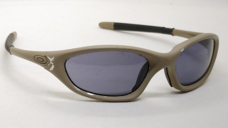 Puede incluir: Gafas de sol color beige con lentes oscuras. Las monturas tienen un dise&ntilde;o curvo y las patillas son negras y beige. El logotipo de Oakley es visible en el lateral de la montura. Las gafas de sol est&aacute;n sobre un fondo blanco.