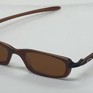 Pode incluir: &Oacute;culos de sol retangulares castanhos com lentes castanhas. As arma&ccedil;&otilde;es e as hastes s&atilde;o de cor castanha lisa. As hastes curvam-se ligeiramente e t&ecirc;m um pequeno detalhe prateado perto da dobradi&ccedil;a.