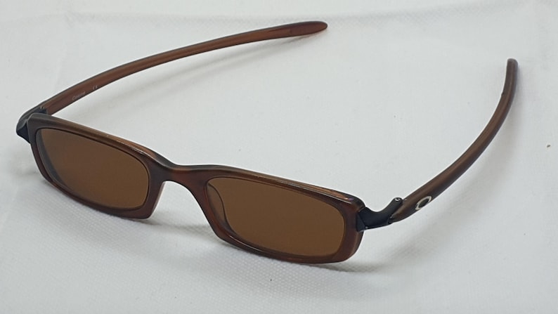 Pode incluir: &Oacute;culos de sol retangulares castanhos com lentes castanhas. As arma&ccedil;&otilde;es e as hastes s&atilde;o de cor castanha lisa. As hastes curvam-se ligeiramente e t&ecirc;m um pequeno detalhe prateado perto da dobradi&ccedil;a.
