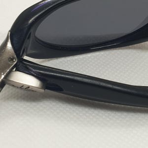 May include: Close-up of black sunglasses with dark lenses. The frame is glossy black, and the hinge is silver. The sunglasses are angled, showing the details of the frame and lens.