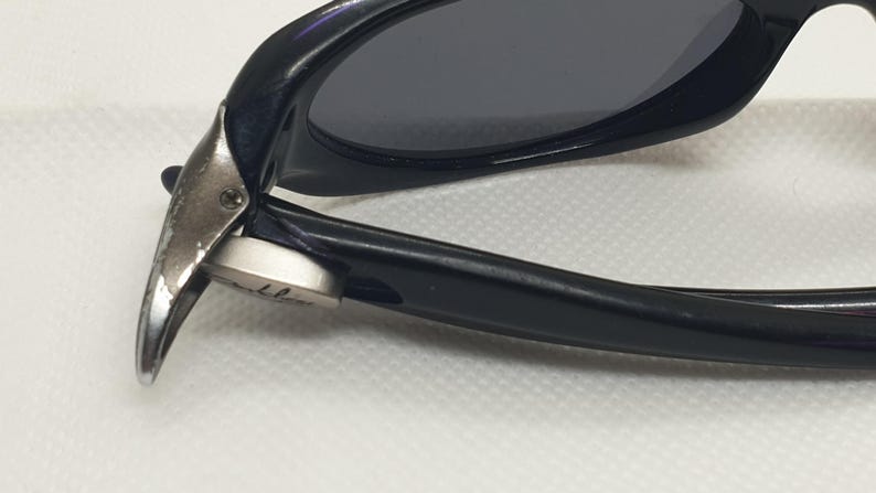 May include: Close-up of black sunglasses with dark lenses. The frame is glossy black, and the hinge is silver. The sunglasses are angled, showing the details of the frame and lens.
