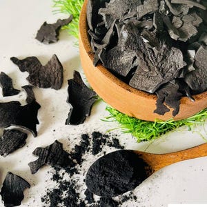 Puede incluir: Un cuenco de madera lleno de trozos de carbón negro y una cuchara de madera con polvo de carbón. La imagen también muestra trozos y polvo de carbón esparcidos sobre una superficie blanca, con hierba verde y el logotipo de Wild Flavours.