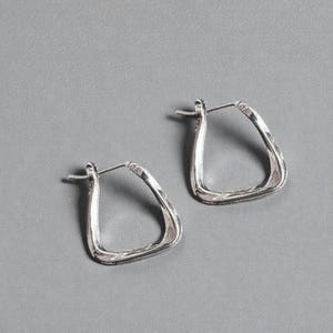 May include: A pair of silver-colored hoop earrings. The earrings have a unique, slightly irregular square shape with a textured surface. The earrings are displayed on a gray background.