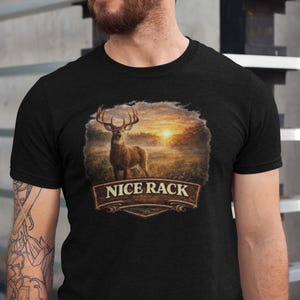 Puede incluir: Camiseta negra con un diseño de ciervo con cuernos grandes sobre un fondo de puesta de sol. El texto "NICE RACK" está debajo. La camiseta es de manga corta.