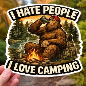 May include: A sticker depicting a Bigfoot character drinking from a bottle, seated by a campfire. The text reads "I HATE PEOPLE" above and "I LOVE CAMPING" below. The background shows a forest and a river.