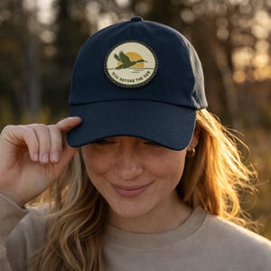 Puede incluir: Gorra de béisbol azul marino con un parche circular. El parche muestra la silueta de un pato volando contra un sol amarillo, con el texto "RISE BEFORE THE SUN". El parche está cosido a la gorra.