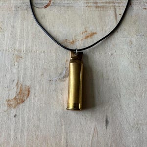 May include: A brass bullet casing pendant hangs from a black cord necklace. The bullet casing is a shiny gold color and is suspended from a small silver-colored loop. The background is a light-colored wooden surface.