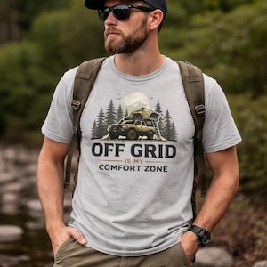 May include: A light gray t-shirt with the text "OFF GRID IS MY COMFORT ZONE" in black. The graphic features a 4x4 truck with a rooftop tent in a forest setting. The person is wearing sunglasses and a backpack.