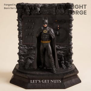 May include: A black Batman statue with a gold belt and bat symbol, standing in front of a dark gray backdrop. The statue is on a black base with the text "LET'S GET NUTS". The backdrop has batarangs and other accessories.