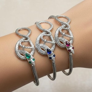 Może przedstawiać: Trzy srebrne bransoletki w kształcie węża z kamieniami szlachetnymi. Jedna ma zielone kamienie, druga niebieskie, a trzecia czerwone. Bransoletki są na nadgarstku, prezentując skomplikowany design i szczegóły biżuterii.