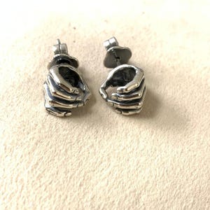 May include: A pair of silver-colored stud earrings. Each earring is shaped like a hand, with detailed fingers and a textured surface. The earrings have a post and butterfly backing for secure wear.