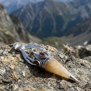 Puede incluir: Un colgante de plata con una piedra de cristal amarilla translúcida en forma de punta. El engaste de metal incluye un intrincado trabajo decorativo en plata y está acentuado con pequeñas piedras de granate rojo oscuro de forma ovalada.