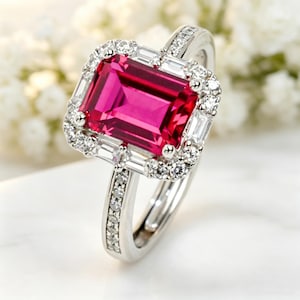 May include: A silver ring featuring a large, rectangular, vibrant pink gemstone, surrounded by a halo of smaller, clear gemstones. The ring's band is also adorned with smaller, clear gemstones, creating a sparkling effect. The ring is set against a soft, blurred background.