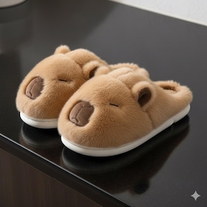 May include: A pair of plush, light brown slippers shaped like capybaras. The slippers have a soft, furry texture, with dark brown accents for the nose, ears, and closed eyes. The soles are white.