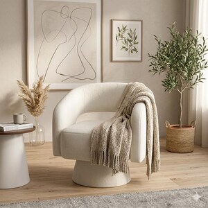 May include: A cream-colored swivel armchair with a beige fringed throw blanket draped over the arm. A small side table with a coffee cup and a vase of dried flowers is to the left. A framed abstract art piece and a framed olive branch print hang on the wall.