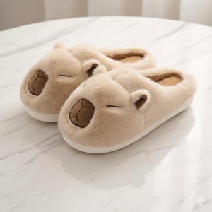 May include: A pair of beige capybara-shaped slippers. The slippers have a soft, plush texture with brown embroidered details for the eyes and nose. The slippers are designed with a closed-toe style and a white sole.