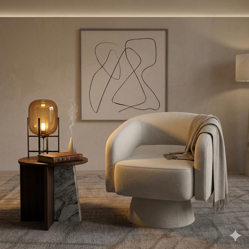May include: A modern living room scene featuring a cream-colored swivel chair with a blanket draped over it. A side table with a lamp and a book sits next to the chair. An abstract art piece hangs on the wall.