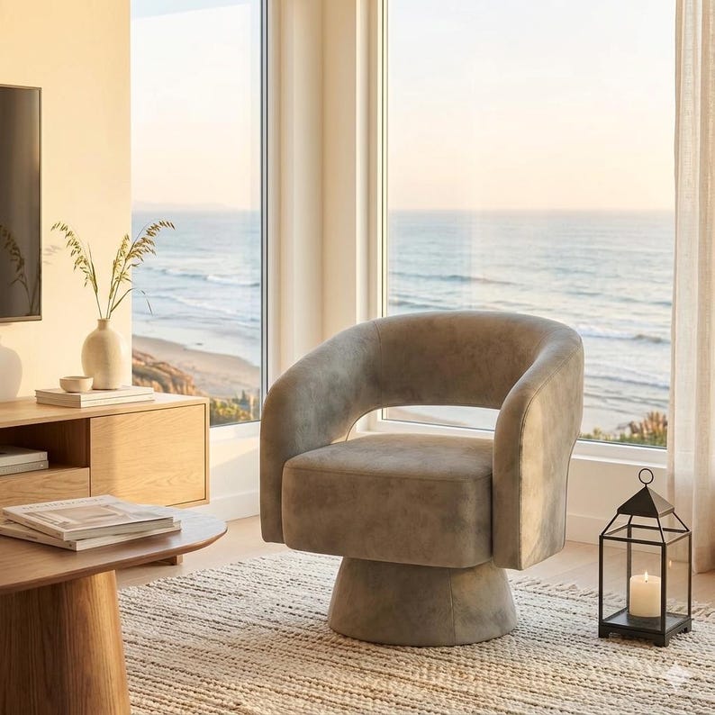 May include: A modern, swivel armchair in a neutral gray hue. The chair features a rounded back and armrests, and a cylindrical base. A black lantern with a lit candle sits nearby. The chair is placed in front of a large window overlooking the ocean.