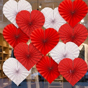 May include: A collection of paper heart decorations in red and white. Some hearts are solid red, while others are white with small red polka dots. The hearts are arranged in a window display, suggesting a festive or celebratory theme.