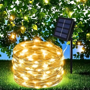Pode incluir: Uma corda enrolada de luzes LED brancas quentes, ideal para decoração exterior, está sobre relva verde. Um pequeno painel solar está posicionado perto, com um fundo de folhas verdes e céu azul. As luzes são projetadas para iluminação eficiente.