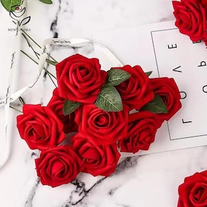 May include: A collection of bright red artificial roses with green leaves. The roses are displayed on a white surface with a marble effect. A white ribbon with text is also visible. The image conveys a floral and decorative theme.