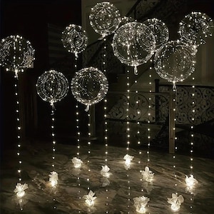 May include: Clear, round balloons filled with tiny white lights, creating a glowing effect. The balloons are suspended from the ceiling and rest on small white paper flower-like bases. The scene is set in a dimly lit room with a staircase in the background.