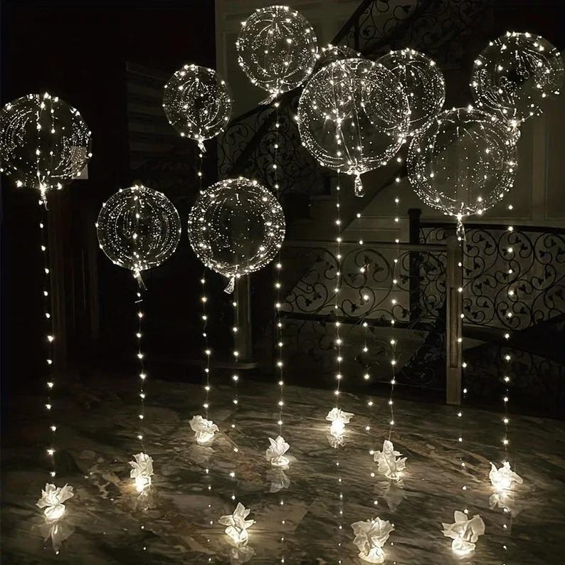 May include: Clear, round balloons filled with tiny white lights, creating a glowing effect. The balloons are suspended from the ceiling and rest on small white paper flower-like bases. The scene is set in a dimly lit room with a staircase in the background.