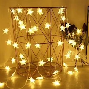 May include: A warm, inviting scene featuring a string of glowing star-shaped lights draped over a geometric wire basket. The lights cast a soft, golden glow, creating a cozy ambiance. The basket is a copper color, and the lights are evenly spaced.