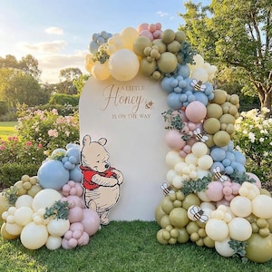 May include: A baby shower display with a Winnie the Pooh theme. The display includes a white arch with the text "A Little Honey Is On The Way", surrounded by pastel balloons and bee cutouts. A Winnie the Pooh cutout is on the left.