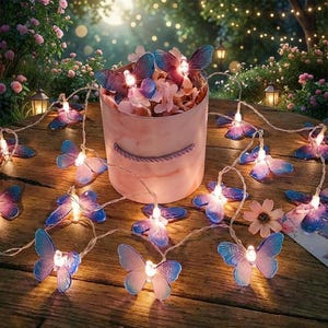 Può includere: Una stringa decorativa di luci a forma di farfalla con ali blu e viola, disposte su un tavolo di legno. Un contenitore rotondo rosa pieno di fiori e altre luci a farfalla è al centro. Lo sfondo mostra un giardino con illuminazione soffusa.