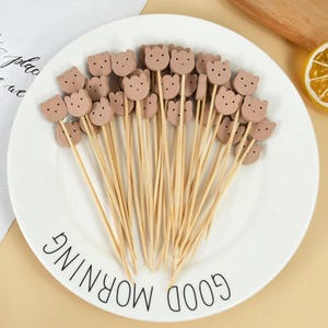 May include: A collection of decorative food picks with bear-shaped toppers. The picks are made of light brown wood, and the bear heads are a matching light brown color with small black dot eyes. The picks are arranged on a white plate with the words "GOOD MORNING" printed on it.