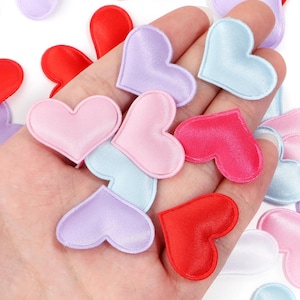May include: A collection of satin heart-shaped appliques in various colors, including red, pink, light blue, and lavender. The hearts are arranged in a hand, showcasing their smooth texture and vibrant hues. Ideal for crafting and embellishments.