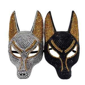 May include: Two animal-shaped masks featuring pointed ears and eye cutouts. The left mask is covered in silver and gold rhinestones, while the right mask is covered in black and gold rhinestones. Both masks have a distinct canine-like facial structure.