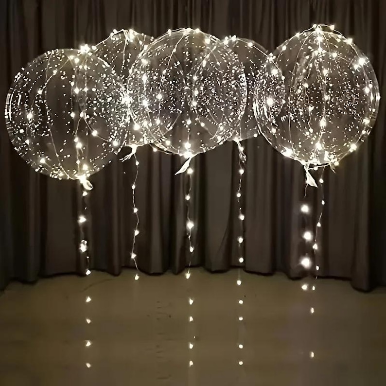 May include: Five clear, round balloons filled with tiny white lights. The lights are evenly distributed throughout the balloons and also hang down from the tied bottoms. The balloons are suspended in front of a dark gray curtain.