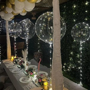 May include: A decorated outdoor dining table with white plates, silverware, and floral centerpieces. Clear balloons with string lights hang above the table. Gold and white balloons are at the top of the image.