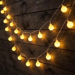 May include: String lights with warm, yellow-toned bulbs are strung against a dark brown wooden background. The bulbs are evenly spaced along a thin, clear wire, creating a soft, inviting glow. The wooden planks have visible grain.