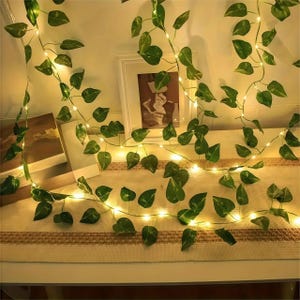 Puede incluir: Una guirnalda de luces decorativa con hojas verdes artificiales. Las pequeñas luces, de tono cálido, crean un brillo suave. Las hojas tienen un aspecto natural, añadiendo un toque de naturaleza a la decoración. La guirnalda está colocada sobre una superficie.